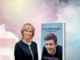 Lesley Roberts next to a copy of her book A Is For Alex on a plae pink cloud background
