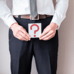 white man in a white shirt, blue trousers, brown belt and grey tie holding a white square with a red question mark on it in front of him, illustrating the question why do men have a foreskin?